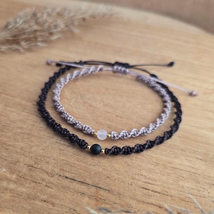 May include: Two macrame bracelets. One is black with a black bead and gold accent, the other is gray with a white bead and gold accent. The bracelets are on a wooden surface.