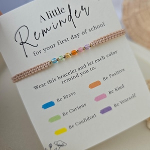Può includere: Un braccialetto in corda intrecciata beige con una fila di piccole perline multicolori blu, verdi, gialle, arancioni, rosa e viola. Il braccialetto è fissato su un cartoncino bianco con il testo A little Reminder for your first day of school.