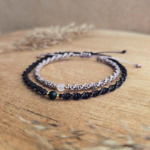 May include: Two macrame bracelets. One is black with a black bead and gold accent. The other is light gray with a clear bead and gold accent. Both bracelets are on a wooden surface.