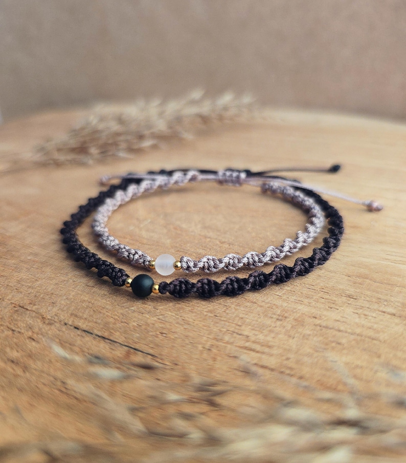 May include: Two macrame bracelets. One is black with a black bead and gold accent. The other is light gray with a clear bead and gold accent. Both bracelets are on a wooden surface.