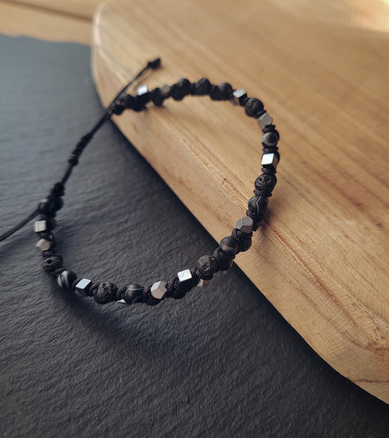 May include: A black and silver beaded bracelet with a black cord closure. The bracelet features black lava rock beads and silver square beads.