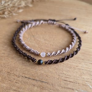 May include: Two macrame bracelets. One is dark brown with a black bead, the other is light brown with a pink bead. Both bracelets have gold-colored accents and are displayed on a wooden surface.