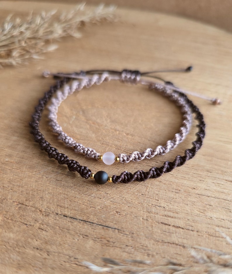 May include: Two macrame bracelets. One is dark brown with a black bead, the other is light brown with a pink bead. Both bracelets have gold-colored accents and are displayed on a wooden surface.