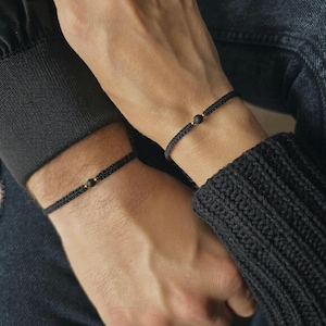 May include: Two black woven bracelets, each with a single dark bead. The bracelets are worn on wrists, with one hand resting on top of the other. The background is blurred, focusing on the bracelets.