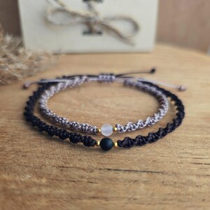 May include: Two handmade macrame bracelets. One is black, the other is gray. Each bracelet has a small, round bead. The bracelets are on a wooden surface, with a gift box in the background.