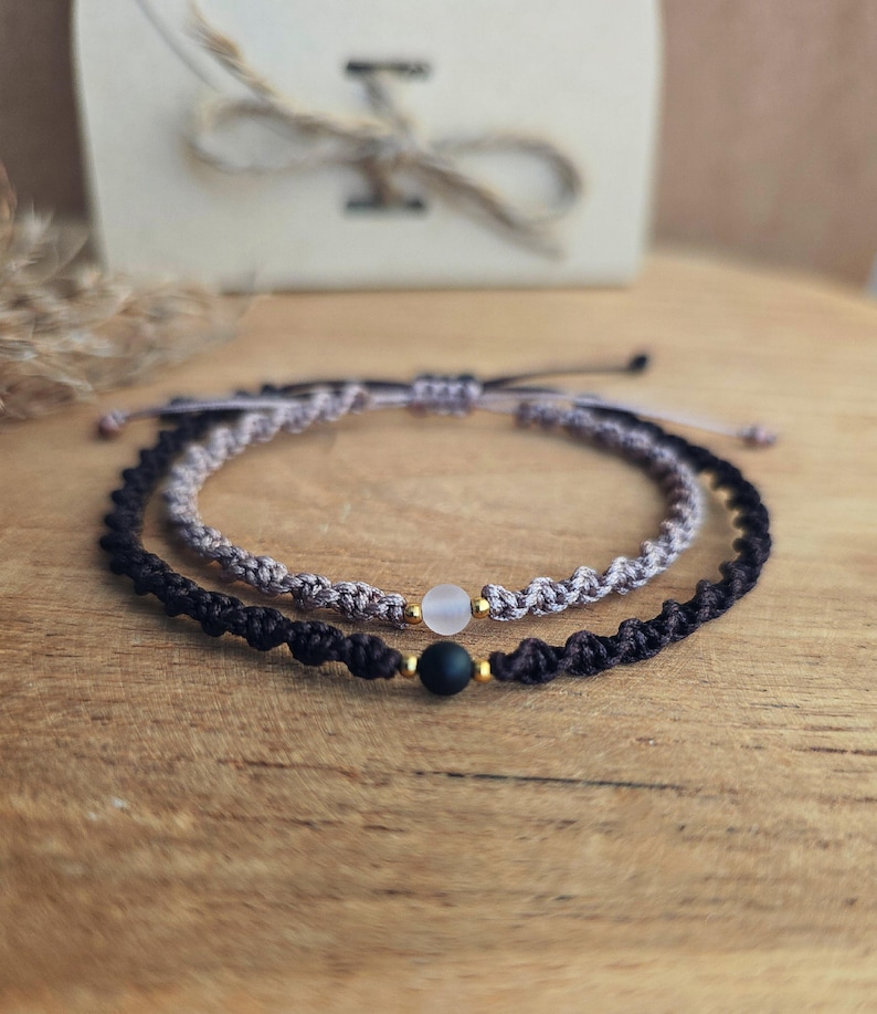 May include: Two handmade macrame bracelets. One is black, the other is gray. Each bracelet has a small, round bead. The bracelets are on a wooden surface, with a gift box in the background.