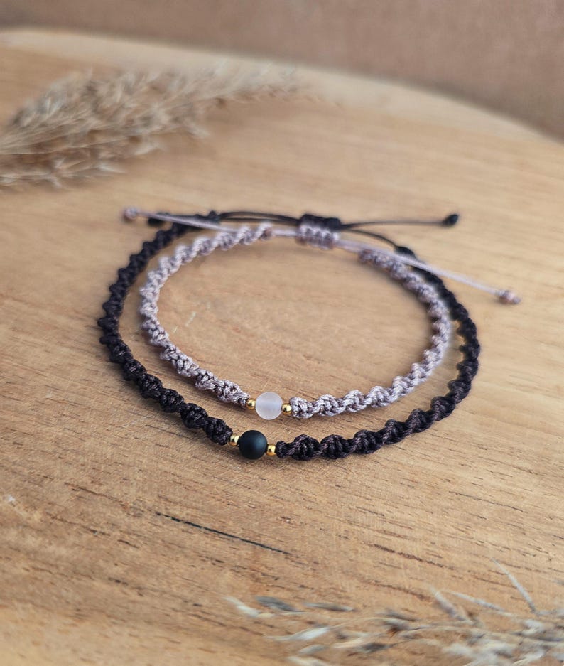 May include: Two macrame bracelets, one black and one gray, each with a small bead. The black bracelet has a black bead, and the gray bracelet has a clear bead. Both bracelets have gold-colored accents and are displayed on a wooden surface.