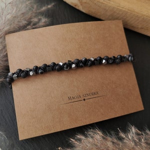 May include: Black beaded bracelet with black lava rock beads and silver metal beads. The bracelet is on a brown paper card with the text "Magia Sznurka".