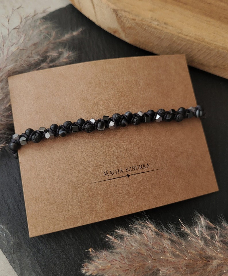 May include: Black beaded bracelet with black lava rock beads and silver metal beads. The bracelet is on a brown paper card with the text "Magia Sznurka".