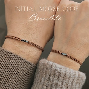 May include: Two brown initial Morse code bracelets are shown. The bracelets are made of woven brown cord with a small, rectangular, dark metal accent. The text "INITIAL MORSE CODE Bracelets" is displayed in white script.