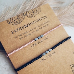 May include: Two bracelets with a message on a card. The card reads "Father & Daughter. To the world, you may be one person, but for me, you are the world. One for you. One for me." The bracelets are black and pink with silver beads.