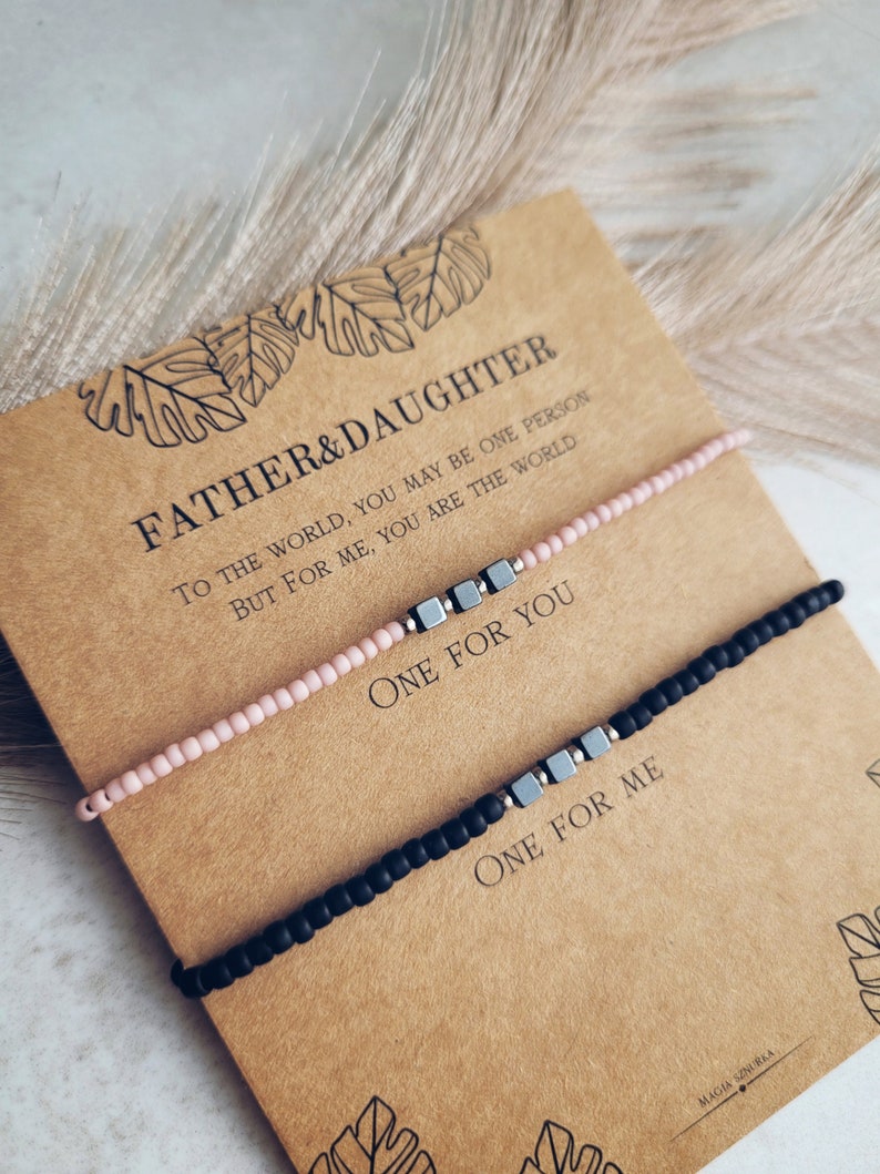 May include: Two bracelets with a message on a card. The card reads "Father & Daughter. To the world, you may be one person, but for me, you are the world. One for you. One for me." The bracelets are black and pink with silver beads.