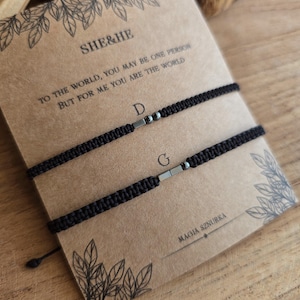 Puede incluir: Dos pulseras negras de macram&eacute; con detalles plateados, presentadas en una tarjeta marr&oacute;n. La tarjeta muestra el texto "SHE&HE" y una cita rom&aacute;ntica. Estas pulseras a juego son ideales para parejas o amigos.