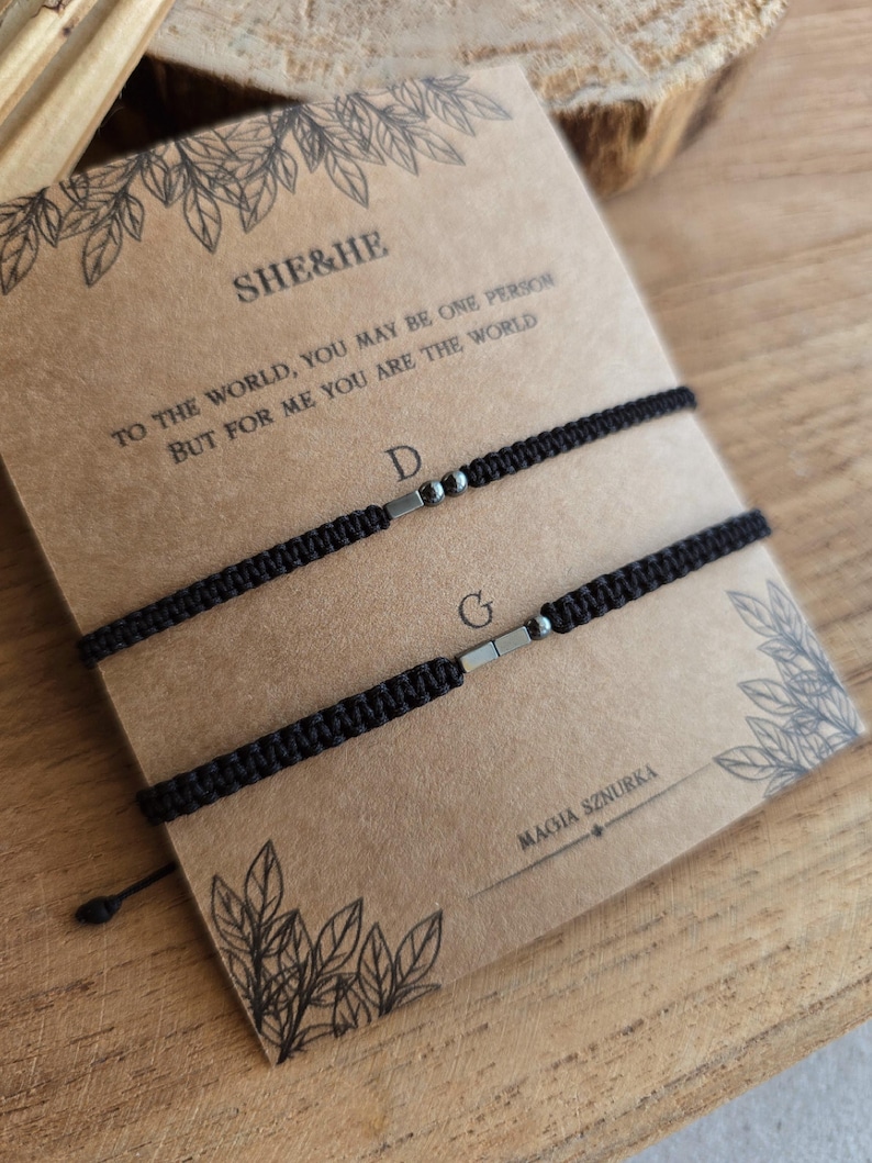 Puede incluir: Dos pulseras negras de macram&eacute; con detalles plateados, presentadas en una tarjeta marr&oacute;n. La tarjeta muestra el texto "SHE&HE" y una cita rom&aacute;ntica. Estas pulseras a juego son ideales para parejas o amigos.