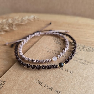May include: Two macrame bracelets, one light brown and one dark brown, each with a small gemstone bead. The bracelets are displayed on a wooden surface with the text "ONE FOR ME" and "ONE FOR YOU".