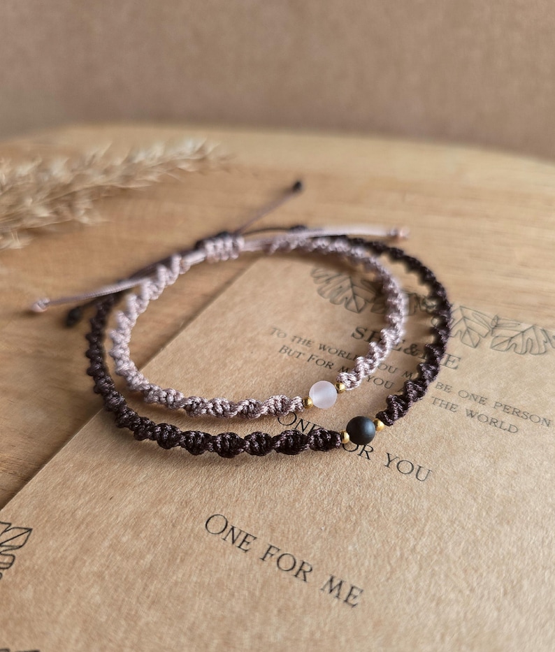 May include: Two macrame bracelets, one light brown and one dark brown, each with a small gemstone bead. The bracelets are displayed on a wooden surface with the text "ONE FOR ME" and "ONE FOR YOU".