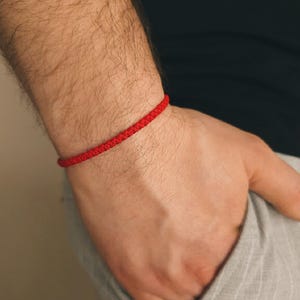 May include: A close-up of a red braided bracelet on a wrist. The bracelet is a vibrant red color and appears to be made of woven threads. The person is wearing a gray pair of pants.