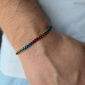 May include: A thin, braided cord bracelet featuring a vibrant rainbow color pattern. The design consists of small, tightly woven segments in red, orange, yellow, green, blue, and purple, interspersed with black thread throughout the entire length.