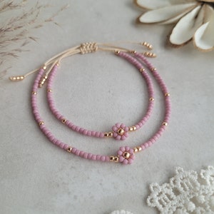 Puede incluir: Dos pulseras delicadas con cuentas rosas y detalles dorados. Cada pulsera presenta un pequeño diseño floral en el centro. Las pulseras son ajustables con un cordón beige.