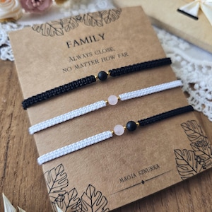 Könnte beinhalten: Drei Makramee-Armbänder, schwarz und weiß, mit Edelsteinperlen, präsentiert auf einer Karte mit der Aufschrift "FAMILY, ALWAYS CLOSE, NO MATTER HOW FAR". Die Karte trägt auch den Text "MAGIA SZURKA".