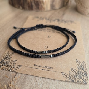 Puede incluir: Dos pulseras negras trenzadas con detalles met&aacute;licos se muestran sobre una tarjeta marr&oacute;n. Las pulseras son ajustables y parecen estar hechas de un cord&oacute;n duradero. La tarjeta tiene el texto "MAGIA SZNURKA" impreso.