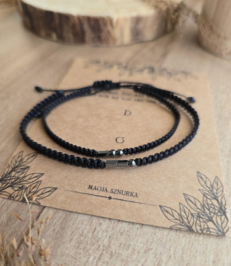 Puede incluir: Dos pulseras negras trenzadas con detalles met&aacute;licos se muestran sobre una tarjeta marr&oacute;n. Las pulseras son ajustables y parecen estar hechas de un cord&oacute;n duradero. La tarjeta tiene el texto "MAGIA SZNURKA" impreso.