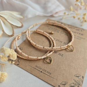 May include: Two delicate bracelets with gold heart charms. The bracelets are made of small, light beige beads and are tied together with a thin, gold cord. The bracelets are displayed on a brown card with the text "To the world you may be one person, but for me you are the world." and "For you" and "Or me" and "Magia Sznurka".