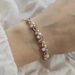 May include: A beaded bracelet featuring a mix of neutral-toned beads in shades of cream, tan, and brown. The bracelet is worn on a wrist, showcasing the delicate design and color palette. The beads are small and round, creating a subtle sparkle.
