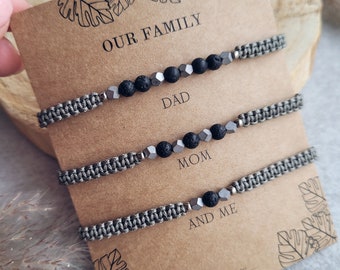 May include: Three gray braided bracelets with black lava beads and silver metal beads. The bracelets are displayed on a brown card with the text "Our Family" at the top and "Dad", "Mom", and "And Me" below each bracelet.