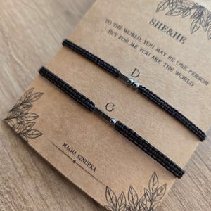 Puede incluir: Dos pulseras negras tejidas con detalles met&aacute;licos plateados se muestran sobre una tarjeta marr&oacute;n. La tarjeta tiene el texto "SHE&HE" y una cita. Tambi&eacute;n son visibles las palabras "MAGIA SZNURKA". El fondo es una superficie de madera marr&oacute;n claro.