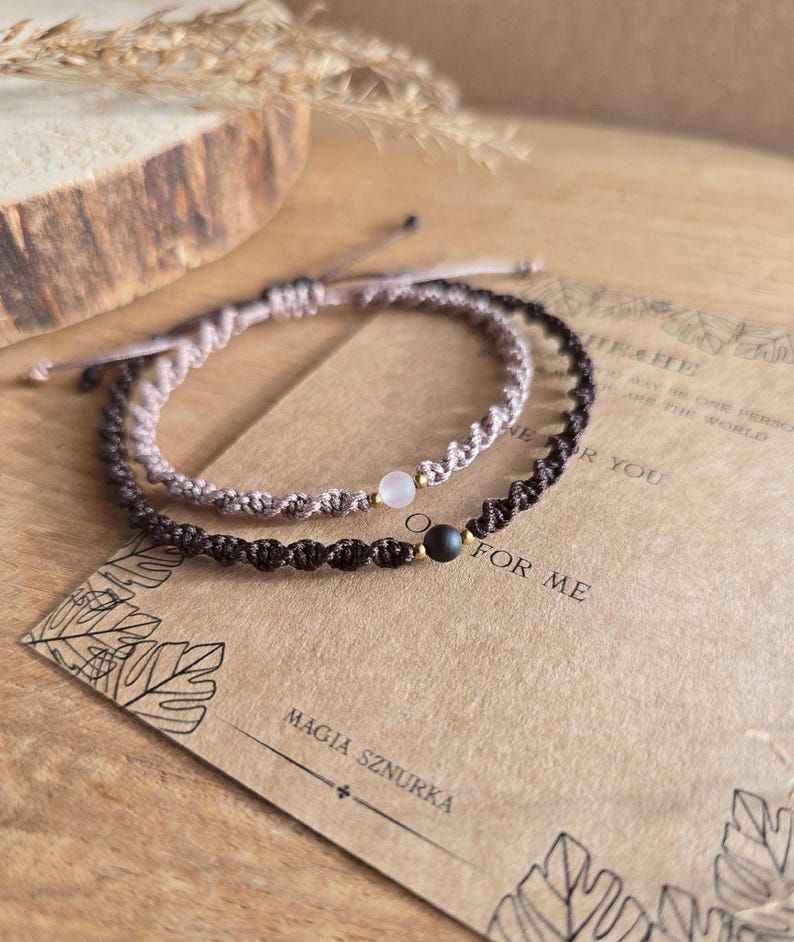 May include: Two macrame bracelets, one light brown and one dark brown, each with a central gemstone bead. The bracelets are displayed on a textured surface with a card that reads "MAGIA SZNURKA" and "FOR ME".