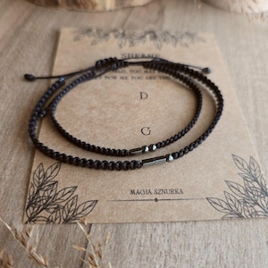 Puede incluir: Dos pulseras negras trenzadas con cordones ajustables, presentadas sobre una tarjeta con texto y dise&ntilde;os florales. La tarjeta dice "SHE&ME" y "MAGIA SZURKA". Las pulseras tienen peque&ntilde;os detalles met&aacute;licos oscuros.