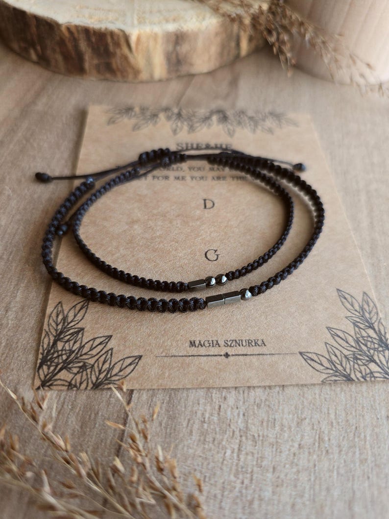 Puede incluir: Dos pulseras negras trenzadas con cordones ajustables, presentadas sobre una tarjeta con texto y dise&ntilde;os florales. La tarjeta dice "SHE&ME" y "MAGIA SZURKA". Las pulseras tienen peque&ntilde;os detalles met&aacute;licos oscuros.