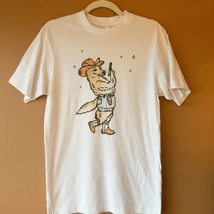 May include: White t-shirt featuring a cartoon illustration of a coyote wearing a cowboy hat and holding a gun. The coyote is surrounded by stars.