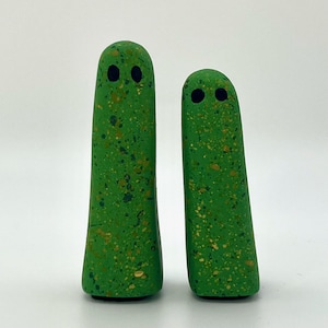 May include: Two green ceramic ghost figurines with black eyes. The ghosts have a speckled green and gold finish.