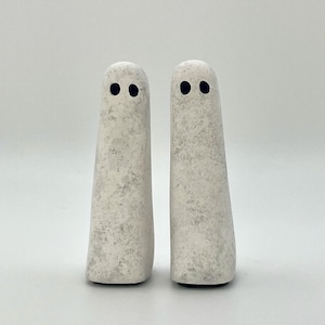 May include: Two white ceramic ghost figurines with black eyes. The ghosts are standing on a white surface.