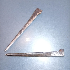 Op de afbeelding: Twee antieke metalen spijkers met een rechthoekige kop en een taps toelopend, puntig lichaam. De spijkers hebben een doffe, verouderde zilverkleur en vertonen tekenen van slijtage. Eén spijker staat verticaal, de andere horizontaal.