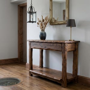 Peut inclure: Une console rectangulaire en bois à la texture brute, dotée de quatre pieds robustes et d'une étagère inférieure. Le plateau supporte un vase en céramique bleu foncé rempli d'herbes séchées et une petite lampe à abat-jour marron foncé.