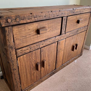 Peut inclure: Un buffet en bois rustique de couleur marron chaud. Il comporte deux tiroirs et deux portes d'armoires, tous avec des poignées rectangulaires simples. La construction du meuble est visible, avec des assemblages et des clous apparents.
