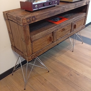 Peut inclure: Meuble TV en bois rustique avec un amplificateur rouge et une platine vinyle sur le dessus. Le meuble a deux tiroirs et repose sur des pieds en épingle à cheveux en métal. Un album de disques est visible dans l'étagère ouverte. Le bois a une finition naturelle.