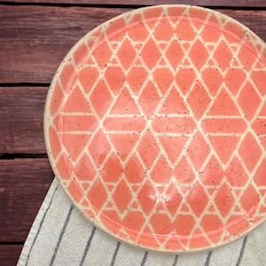 May include: A ceramic plate with a pink and white geometric pattern. The plate has a white rim and a pink center with a white diamond pattern.