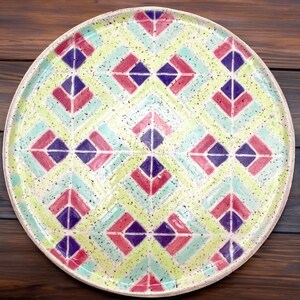 May include: A round ceramic plate with a geometric pattern in shades of pink, purple, green, and white. The plate has a speckled glaze and a white background.