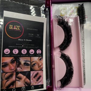 Puede incluir: Una caja de plástico rosa y transparente que contiene dos pares de pestañas postizas negras y un cepillo aplicador de pestañas negro. La caja tiene una etiqueta que dice "Blaze N Minks" e incluye una dirección de sitio web y un número de teléfono.