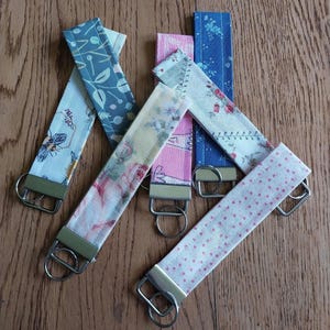 Op de afbeelding: Vijf stoffen sleutelhangers met verschillende patronen en kleuren. De sleutelhangers zijn bevestigd aan zilveren sleutelringen. De patronen omvatten bloemen, stippen en geometrische ontwerpen.