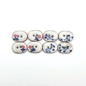 May include: Eight white oval buttons with floral designs in pink and blue. The buttons have two holes each and a brown border. The floral patterns include small flowers and leafy branches, creating a delicate, handcrafted look.