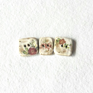 May include: Three rectangular, off-white buttons with floral designs. Each button features a different arrangement of red flowers and green leaves. The buttons have two holes for sewing and a slightly distressed, vintage appearance.