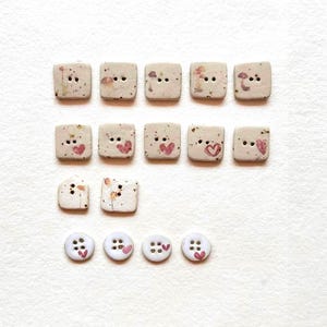 May include: A collection of decorative buttons in various shapes and sizes. The square buttons feature painted designs of hearts and mushrooms. The round buttons are white with pink heart accents. The buttons are ideal for sewing and craft projects.
