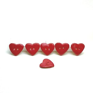 Puede incluir: Seis botones rojos en forma de corazón. Cinco botones están dispuestos en fila, con un botón debajo. Cada botón tiene dos agujeros para coser. Los botones son de color rojo brillante.