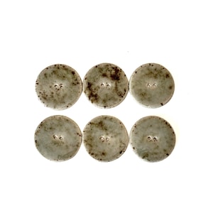May include: Six round, weathered-looking buttons with two holes each. The buttons are a muted gray-green color with darker brown speckling, giving them a vintage or antique appearance. They are arranged in two rows of three.