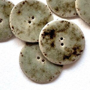 May include: Close-up of several round, light green buttons with two holes each. The buttons have a speckled, mottled appearance with dark brown spots around the edges, giving them a rustic, vintage look. These buttons are ideal for sewing and craft projects.